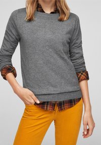 Frau in Schichten gekleidet, trägt einen grauen Pullover über einem orangefarbenen karierten Hemd, kombiniert mit senfgelben Hosen, die Hand in der Tasche.