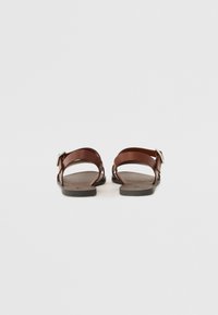 Brune lær sandaler med åben hæl, der har to krydsede stropper og en metal spænde. Sålen er flad med en tekstureret overflade.