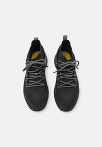 Paire de baskets en maille noire avec des lacets gris et des semelles intérieures jaunes, vues de dessus sur un fond blanc.
