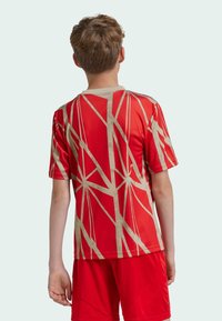 Camisa roja de manga corta con un patrón geométrico en beige. El tejido parece suave y deportivo. Vista trasera que muestra el cuello redondo y la continuidad del diseño.