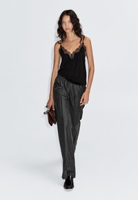 Zwarte camisole met kantafwerking, gestyled met grijs gestreepte broek. Het model draagt een bruine clutch en zwarte schoenen, staand tegen een witte achtergrond.