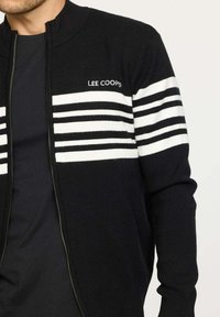 Gilet noir avec des rayures horizontales blanches sur la poitrine. Présente une fermeture éclair et un col haut. Logo "LEE COOPER" sur le côté gauche.