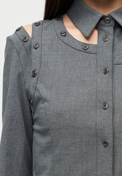 Γκρι πουκάμισο με κουμπιά, διαθέτει γιακά, ανοίγματα στους ώμους και μαύρα κουμπιά κατά μήκος του ώμου για πρόσθετη λεπτομέρεια και σχεδιασμό.