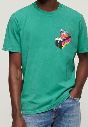 Mężczyzna w zielonym t-shircie z kolorową grafiką czerwonego vana i desek surfingowych na piersi, w połączeniu z niebieskimi dżinsami.