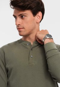 Junger Mann in olivgrünem Henley-Shirt, der nach links schaut, mit silberner Armbanduhr am gehobenen rechten Handgelenk vor einem schlichten Hintergrund.