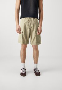 Pantaloni cargo beige con due tasche laterali, tessuto leggero e vestibilità comoda, abbinati a un top senza maniche nero e scarpe da ginnastica marroni.