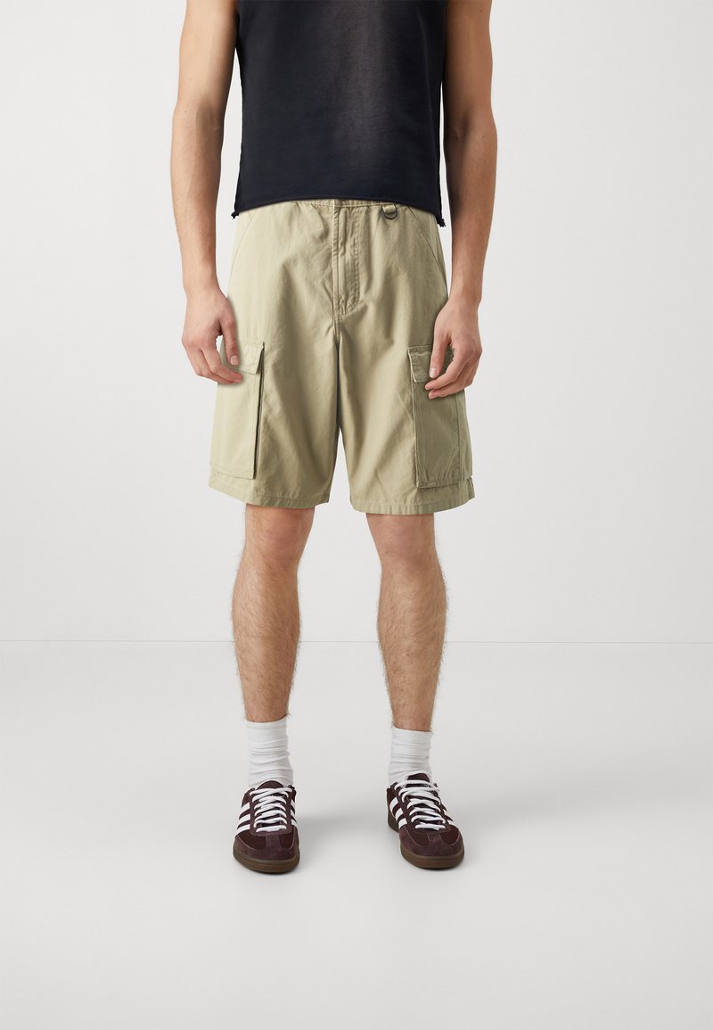 Pantaloni cargo beige con due tasche laterali, tessuto leggero e vestibilità comoda, abbinati a un top senza maniche nero e scarpe da ginnastica marroni.