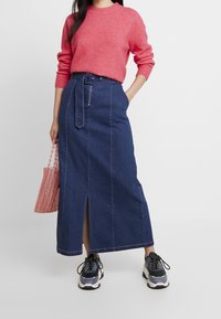 Denim maxikjol med sprund fram, mörkblå färg och midja med bälte. Kombinationen med en rosa texturerad tröja och svarta sportiga sneakers.