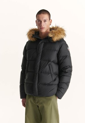 Zwarte pufferjas met een afneembare capuchon, voorzien van een faux fur rand, diagonale stiksels en zijzakken. Draagt over olijfgroene broeken.