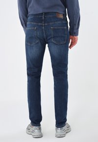 Bărbat îmbrăcat în blugi negri, pantofi sport gri și o cămașă bleumarin cu nasturi, stând cu spatele la cameră.