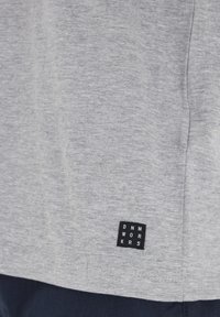 T-shirt grigio a maniche corte realizzato in tessuto misto cotone con una texture morbida, caratterizzato da un'etichetta nera con testo bianco in basso a sinistra.