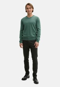 Pulover verde cu decolteu în V dintr-un material tricotat moale, asortat cu pantaloni închiși la culoare și încălțăminte neagră, având o siluetă ajustată și manșete cu nervuri.