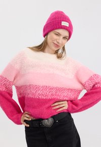 Rosa Strickpullover mit Farbverlaufstreifen, die von hellem Rosa bis leuchtendem Fuchsia reichen. Ein Paar rosa gerippte Mütze mit einem Etikett. Schwarzer Gürtel.