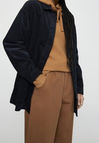 Chaqueta de camisa de pana azul marino con cierre de botones, combinada con una sudadera color beige y pantalones marrones. Tejido texturizado y corte relajado.