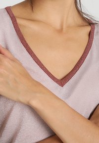 Lyserød V-hals top med en glitrende tekstur og en mørkere maroon kant omkring halsudskæringen, close-up visning af stoffet og armen.