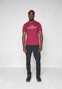 Burgundowa bawełniana koszulka z białym napisem "Peak Performance", w połączeniu z czarnymi lekkimi spodniami i czarnymi butami trekkingowymi. Prosty, funkcjonalny design.