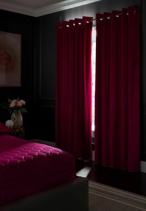 Camera da letto scura con tende rosso intenso parzialmente aperte, copriletto rosso trapuntato abbinato, un comodino con un vaso fiorito e una lampada moderna.