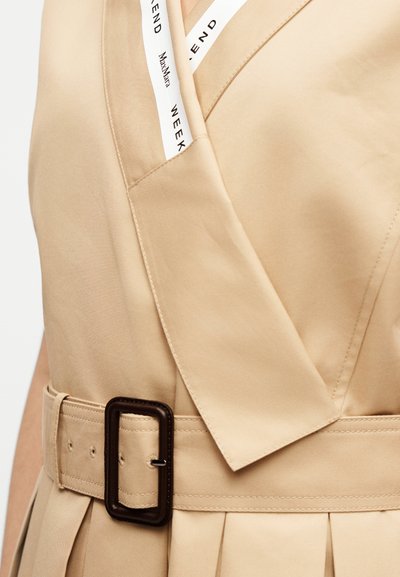 Μπεζ παλτό βροχής με φαρδιούς γιακάδες και μία ζώνη που ταιριάζει, με ένα σκούρο ορθογώνιο πόμολο, κοντινή λήψη στην περιοχή της μέσης και του στήθους.