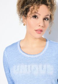 Světle modré tričko s dlouhým rukávem s nápisem "UNIQUE" vyšitým texturálními písmeny na přední straně. Má široký výstřih a měkkou tkaninu.