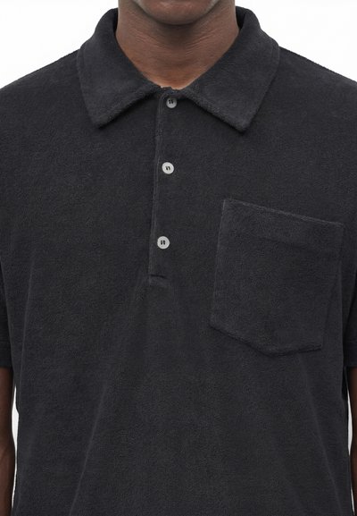 Άνδρας που φοράει μαύρο polo από πετσέτα με κοντά μανίκια, γιακά, τρία κουμπιά και τσέπη στο στήθος, εμφανιζόμενος από τον λαιμό έως τη μέση.