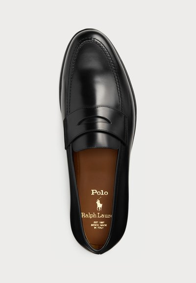Mocasín de cuero negro con un acabado suave, que presenta una correa decorativa, punta redondeada y un logotipo dorado en la plantilla marrón.