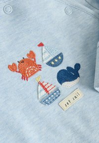 Tissu bleu clair avec un crabe orange brodé, deux voiliers, une baleine bleue et une étiquette « prendre la mer ».