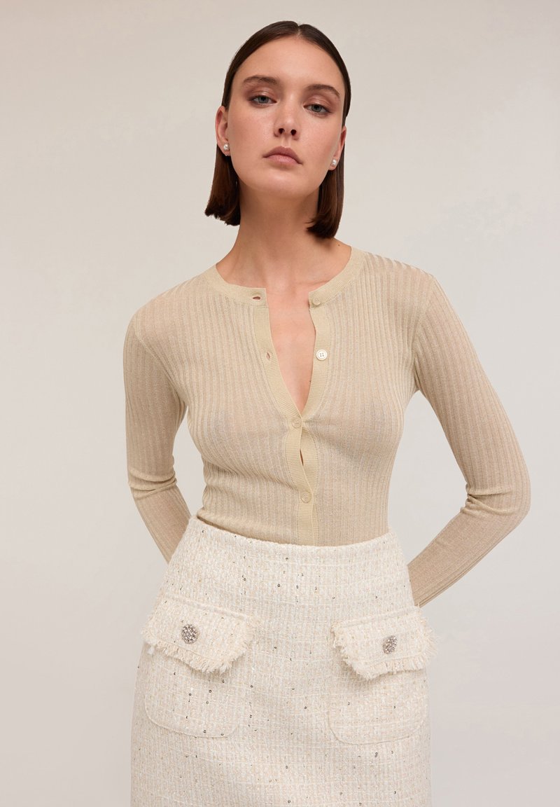 Cardigan bej cu dungi, cu mâneci lungi și închidere cu nasturi, asociat cu o fustă albă texturată ce are două buzunare frontale și nasturi decorativi.