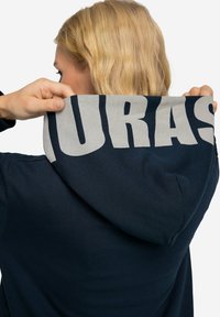 Marinblå huvtröja med en stor grå grafikkon på huvan. Materialet är mjukt med en slät textur. Synlig sömnadsdetalj vid axlarna.