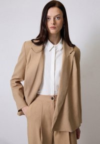 Bežna blazer jakna iz mehkega materiala, z zareznimi reverji in odprtim sprednjim delom. Kombinirana z belo srajco s ovratnikom. Prilagojena kroja.