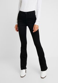 Femme portant un pantalon bootcut noir ajusté, un haut blanc à manches longues et des bottines blanches à talons pointus, debout sur un fond uni.