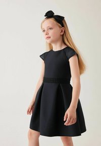 Schwarzes Kleid mit kurzen, durchsichtigen Ärmeln, tailliertem Oberteil und ausgestelltem Rock. Mit einem strukturierten schwarzen Gürtel. Kombiniert mit einem schwarzen Haarband mit Schleife.