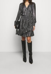 Robe noire à manches longues avec un design portefeuille, présentant un motif texturé et un ourlet à volants. Associée à des bottes en cuir noires montantes jusqu'aux genoux et un sac à main.