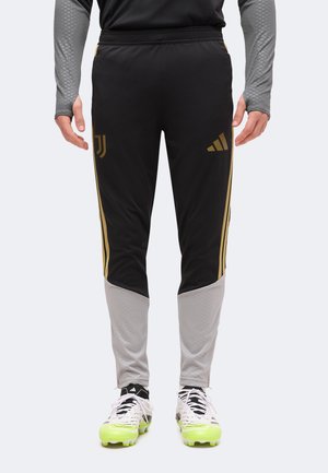 Partie inférieure du corps d'une personne portant un pantalon de sport noir et gris avec des bandes dorées et des logos, ainsi que des crampons blancs avec des semelles vert néon.