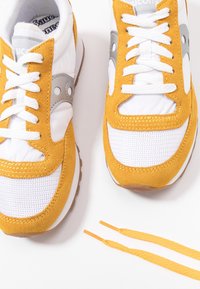 Paire de baskets Saucony blanches et jaune moutarde avec lacets blancs, semelles brunes et lacets jaunes moutarde supplémentaires à côté.