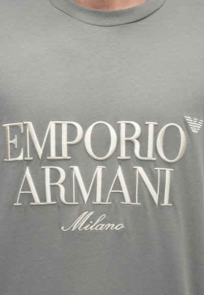 T-shirt cinza de algodão com texto bordado em branco "EMPORIO ARMANI Milano" e um pequeno detalhe de logótipo no canto superior direito.