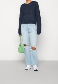 Pull en maille bleu marine, jean bleu clair déchiré, baskets blanches et sac à main en maille verte. Coupe décontractée, col rond et détails de couture visibles.
