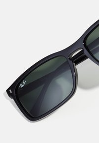 Ochelari de soare din plastic negru cu lentile verzi. Formă pătrată și rame largi. Logo-ul Ray-Ban este vizibil pe lentila stângă. Bentițe subțiri cu un finisaj lucios.
