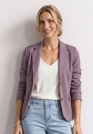 Femme souriante aux cheveux blonds en chignon bas, portant un blazer mauve, une blouse blanche, un jean bleu clair et un bracelet argenté, debout devant un mur clair.