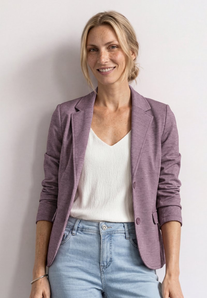 Femme souriante aux cheveux blonds en chignon bas, portant un blazer mauve, une blouse blanche, un jean bleu clair et un bracelet argenté, debout devant un mur clair.