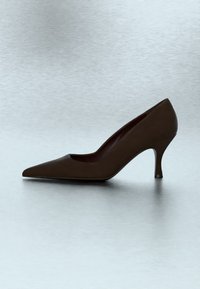 Chaussure à talon haut pour femme couleur marron foncé à bout pointu avec un talon fin de hauteur moyenne sur une surface gris clair.