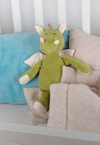 Green plush dragon toy with beige accents sits among beige and blue pillows and blankets, featuring wings and a textured fabric.