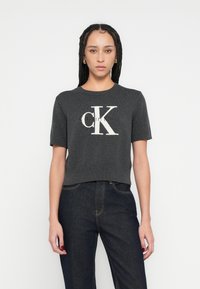 Tricou scurt gri tricotat, cu mâneci scurte, având un logo mare alb „CK” pe față, cu decolteu și tiv striați.
