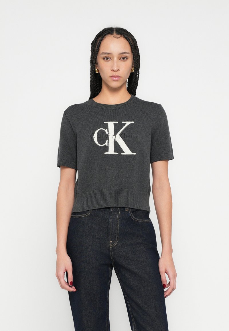 Tricou scurt gri tricotat, cu mâneci scurte, având un logo mare alb „CK” pe față, cu decolteu și tiv striați.