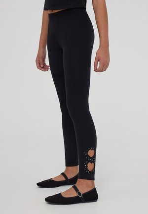 Leggings neri in tessuto elastico, con forme a cuore ritagliate e borchie decorative all'orlo. Indossati con scarpe nere basse.