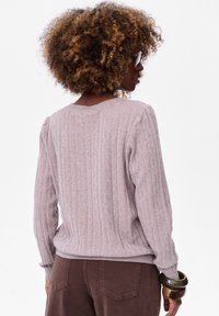 Lichtgrijze geribbelde trui met een ronde halslijn en lange mouwen, met subtiele textuurdetails. Gecombineerd met een paarse broek en een groene armband.