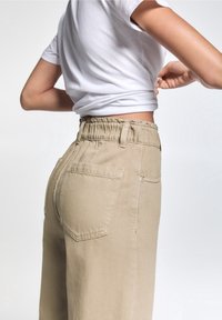 Pantalones beige de cintura alta con una cinturilla elástica, con bolsillos traseros y un tejido texturizado. Combinados con una camisa blanca de manga corta.