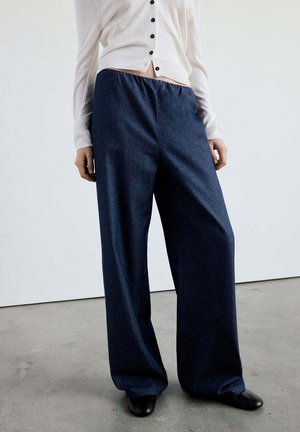 Persona que lleva pantalones azules oscuros de pierna ancha, camiseta blanca con botones y zapatos negros, de pie sobre suelo gris frente a pared blanca lisa.