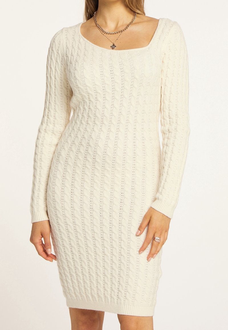 Femme portant une robe-pull ajustée à manches longues, en maille torsadée crème, avec un décolleté carré et un collier chaîne avec pendentif.