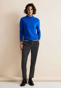 Maglione blu a maglia con colletto alto, abbinato a pantaloni grigio scuro con un motivo testurizzato. Scarpe nere stringate completano l'outfit.