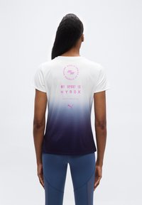 Γυναίκα που φοράει μπλούζα με ομπρέ από λευκό σε ναυτικό μπλε και ροζ κείμενο "MY SPORT IS HYROX" στην πλάτη, συνδυασμένη με μπλε αθλητικό κολάν.